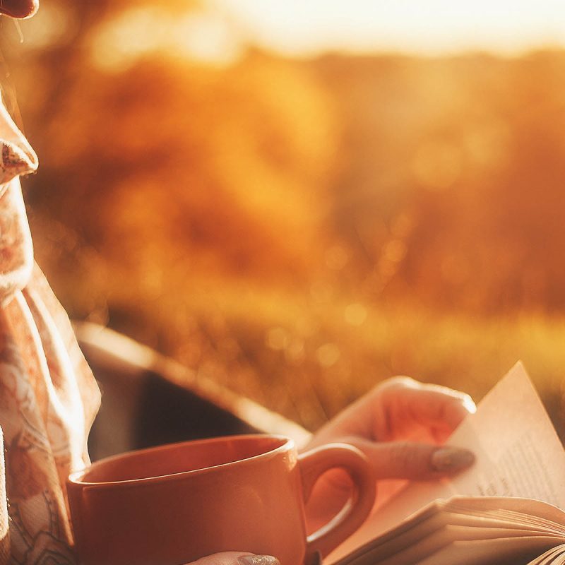 A woman relaxing in the warm Autumn landscape reading a book and drinking a hot drink